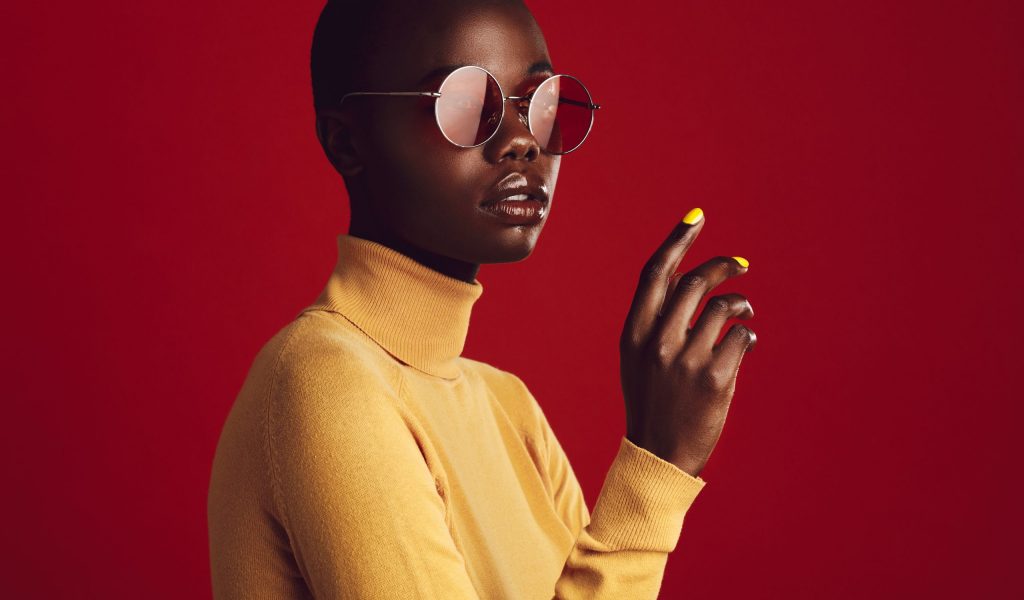 Close up portrait of african female with buzz cut hairstyle against red background. Beautiful african woman in casuals wearing sunglasses looking at camera.