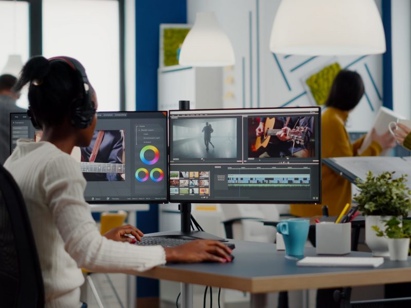 Black videographer putting on headset editing movie using post production software working in creative start up agency office. African editor processing video footage on computer with dual displays