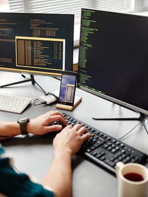 Closeup of IT developer typing on keyboard with programming code on computer screen while working in office interior, copy space